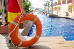 Close up of a lifebuoy ring with rope held up by a lifeguard with a swimming pool on the background . Lifeguard during work concept