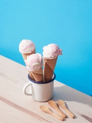 Three strawberry ice cream cones inside a white cup on a blue background with wooden spoons on a wooden table. Harsh light with shadows.