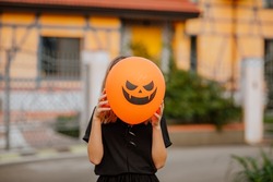 Young teen girl in black clothes standing against orange house and holding halloween balloon before her face . Holiday concept. 