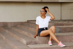 An African girl in a white T-shirt and glasses sits on the steps. Summer