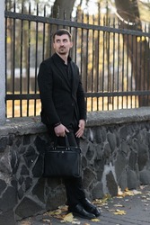 A confident young bearded man in a black suit and case is waiting for a meeting with a colleague to resolve work issues. Working meeting on the street