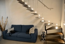 Decorative stairs in the apartment made of bronze material. Above them is a light from lamps. A sofa with pillows under the stairs. The concept of a modern apartment