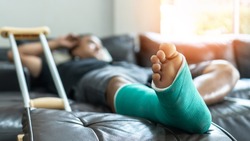 Bone fracture foot and leg on male patient with splint cast and crutches during surgery rehabilitation and orthopaedic recovery lying on couch staying at home