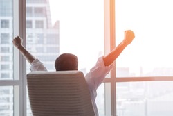Business achievement concept with happy  businessman relaxing at work in office room, resting and raising fists with ambition success looking forward to city building urban scene through glass window