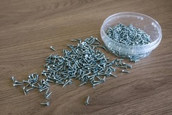 Small silver screws are lying on a wooden table.Metal fasteners.A box with chrome cogs.