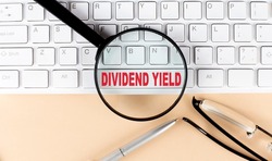 Text DIVIDEND YIELD on a keyboard with magnifier , glasses and pen on beige background