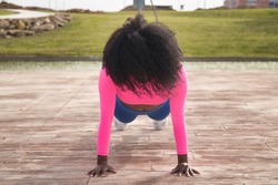 African-American woman with afro hair and sportswear, with fluorescent pink t-shirt and leggings, doing push-ups next to an outdoor lake. Fitness concept, sport, street, urban.