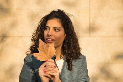 Portrait of young and beautiful woman, brunette, with curly hair with jacket, holding a dry leaf with her hands in front of her face and innocent look. Concept beauty, fashion, autumn, innocence.