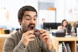Hispanic man with mustache eating a chocolate sweet bun. Unhealthy habits in the office concept.