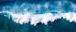A forceful, crashing wave of the ocean is captured in this stunning aerial photo, with the massive wave breaking into a white spray against the deep waters below, evoking feelings of awe and power.