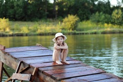 Little girl in a straw hat and swimsuit at a river lake shore on sunset sitteing on wooden pier. Relaxing in nature