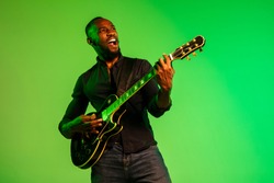 Young african-american musician playing the guitar like a rockstar on gradient green-yellow background. Concept of music, hobby, festival, open-air. Joyful attractive guy improvising, singing song.
