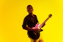 Young african-american musician playing the guitar like a rockstar on yellow background in neon light. Concept of music, hobby, festival, open-air. Joyful attractive guy improvising, singing song.