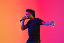 Young african-american musician singing on gradient orange-purple studio background in neon light. Concept of music, hobby, festival. Joyful party host, stand upper. Colorful portrait of artist.
