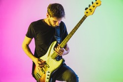Close up young and joyful caucasian musician playing bass guitar on gradient studio background in neon light. Concept of music, hobby, festival. Colorful portrait of modern artist. Inspired