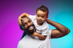 African-american father and son portrait on gradient studio background in neon. Beautiful male models in casual style, white shirt. Concept of human emotions, facial expression