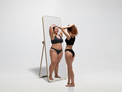 Young fit, slim woman looking at fat girl in mirror's reflection on white background. Thinking she's not enough sportive. Concept of healthy lifestyle, fitness, sport, nutrition and body positive.