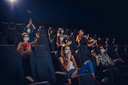 Cinema in quarantine. Coronavirus pandemic safety rules, social distance during movie watching. Men, women in protective face mask sitting in a rows of auditorium. Leisure time, youth culture concept.