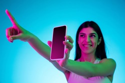 Showing screen, pointing. Brunette woman's portrait on blue studio background in mixed neon. Beautiful model with smartphone. Concept of human emotions, facial expression, sales, ad, fashion