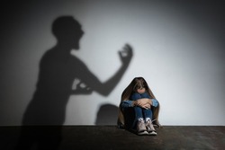 Domestic violence, abusing. Scared little caucasian girl, victim sitting close to white wall with shadow of angry father's threatening on it. Awareness of social problem, childhood, physical violence.