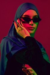 Style. Beautiful arab woman in stylish hijab isolated on burgundy studio background in neon light with copyspace for ad. Fashion, beauty, style concept. Female model with trendy make up, accessories.
