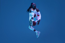 Flying. Beautiful african-american female basketball player in motion and action in neon light on blue background. Concept of healthy lifestyle, professional sport, hobby. Woman in sport.