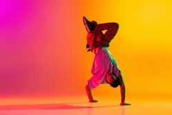 Headstand. Portrait of young stylish man, break dancing dancer training in casual clothes isolated over gradient pink yellow background. Youth culture, movement, street style and fashion, action.