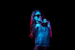 Young happy girl in sunglasses shopping bags, cheerful. Portrait of young woman in neon light on dark backgound. The human emotions, black friday, cyber monday, purchases, sales, finance concept.