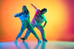Dancing couple. Stylish man and woman dancing hip-hop in casual youth clothes on gradient yellow red background at dance hall in neon light. Youth culture, hip-hop, fashion, breakdance, action and ad