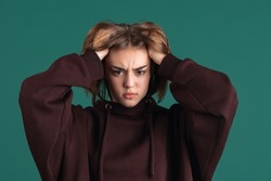 Angry, annoyed. Portrait of young beuatiful girl, student in brown hoodie has a headache isolated on green studio backgroud. Human emotions, facial expression, youth, fashion, ad concept.