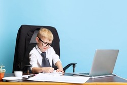 Hard project. One little boy, child businessman in huge jacket suit sitting at office table isolated on blue studio background. Concept of smart childhood, study, games, career, imitation, emotions.