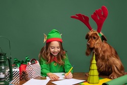Funny friend. Little beautiful kid dressed like funny gnome or elf sitting at table with her funny dog and writing letter to Santa Claus. Merry Christmas and New Year concept. Looks happy