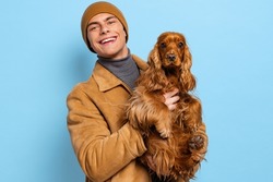 Young handsome happy man, student wearing warm winter clothes and hat standing with cute dog on blue studio background. Concept of human emotions, facial expression, study, ad, fashion, beauty.