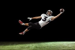 Flying, catching ball. Portrait of American football player training isolated on dark studio background with green grass flooring. Concept of sport, movement, achievements. Copy space for ad