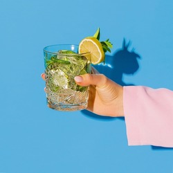 Exclusive alcoholic drink. Female hand holding glass with cocktail mojito with lemon isolated on light blue neon background. Complementary colors, blue, yellow and green. Copy space for ad, design