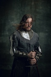 Half-length portrait of brutal seriuos man in image of medieval warrior or knight with dirty wounded face holding big sword isolated on dark background. Comparison of eras, history, renaissance style