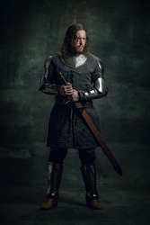 Vintage style portrait of brutal seriuos man, medieval warrior or knight with dirty wounded face holding big sword isolated over dark background. Comparison of eras, history, renaissance style