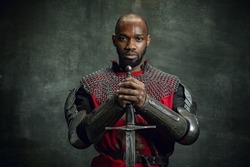 Courage. Vintage portrait of dark skinned medieval warrior wearing chain mail holding big sword isolated over dark vintage background. Comparison of eras, history, renaissance style. Human emotions