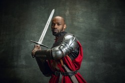 Attack. Vintage portrait of dark skinned medieval warrior wearing chain mail holding big sword isolated over dark vintage background. Comparison of eras, history, renaissance style. Human emotions