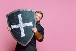 Portrait of scary man, medieval warrior or archer in armor hiding himself behind a shield isolated over pink studio background. Frightened fighter. Comparison of eras, history, renaissance style