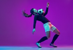 Emotive girl, female soccer, football player in motion, action isolated on purple studio background in neon light. Concept of sport, action, motion, fitness, team, achievements and goals