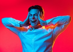 Half-length portrait of young man, student in white shirt toughing head and screaming isolated over red studio background in blue neon light. Concept of youth, emotions, facial expression