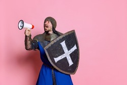 Portrait of man, medieval warrior or knight in armor with shield shouting in megaphone isolated over pink studio background. Wounded face. Comparison of eras, history, renaissance style
