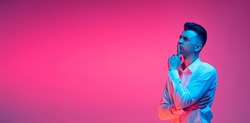 Ideas. Flyer with portrait of young handsome man in white shirt thinking, praying isolated over magenta color studio background in blue neon light. Concept of youth, fashion, emotions, facial