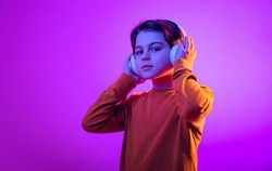 Portrait of boy, child listening to music in headphones, posing isolated over purple background in neon. Music lover. Concept of emotions, childhood, facial expression. Copy space for ad