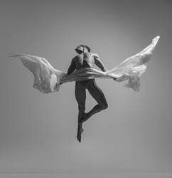 Flight. Black and white portrait of graceful muscled male ballet dancer flying with fabric, cloth isolated on grey studio background. Grace, art, beauty, contemp dance concept. Weightless