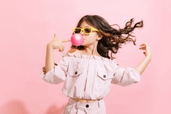 Happy and carefree childhood. Fashionable little girl, kid blowing gum bubble isolated over pink color background. Concept of children emotions, fashion, beauty, back to school, travel and ad