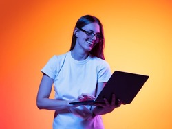 Remote studying. Portrait of young smiling girl, student in white t-shirt with laptop isolated on orange color background in neon light. Concept of beauty, art, fashion, education