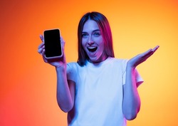 One astonished glad woman with in white t-shirt showing screen of smart phone in hand with amazed expression isolated on orange background in neon. Online shopping sale concept