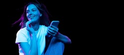 Online shopping. Happy young pretty girl with cellphone isolated on dark background in purple neon light. Concept of emotions, facial expression, youth, aspiration.